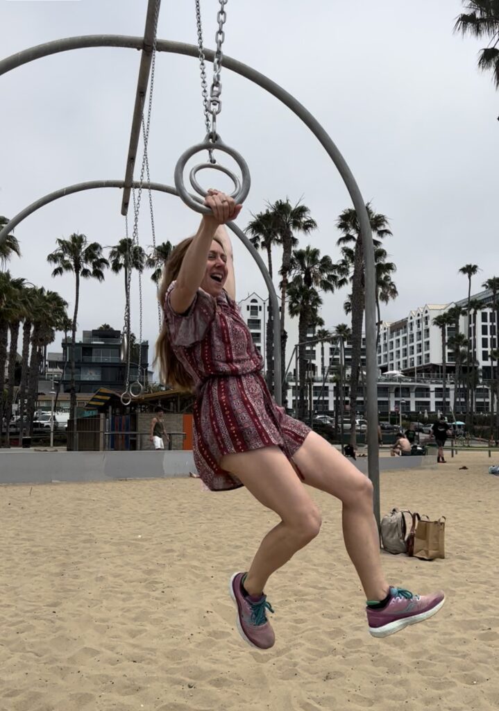 woman swinging on hanging monkey bar rings at beach. Wearing a red shorts and top and pink gym shoes.
